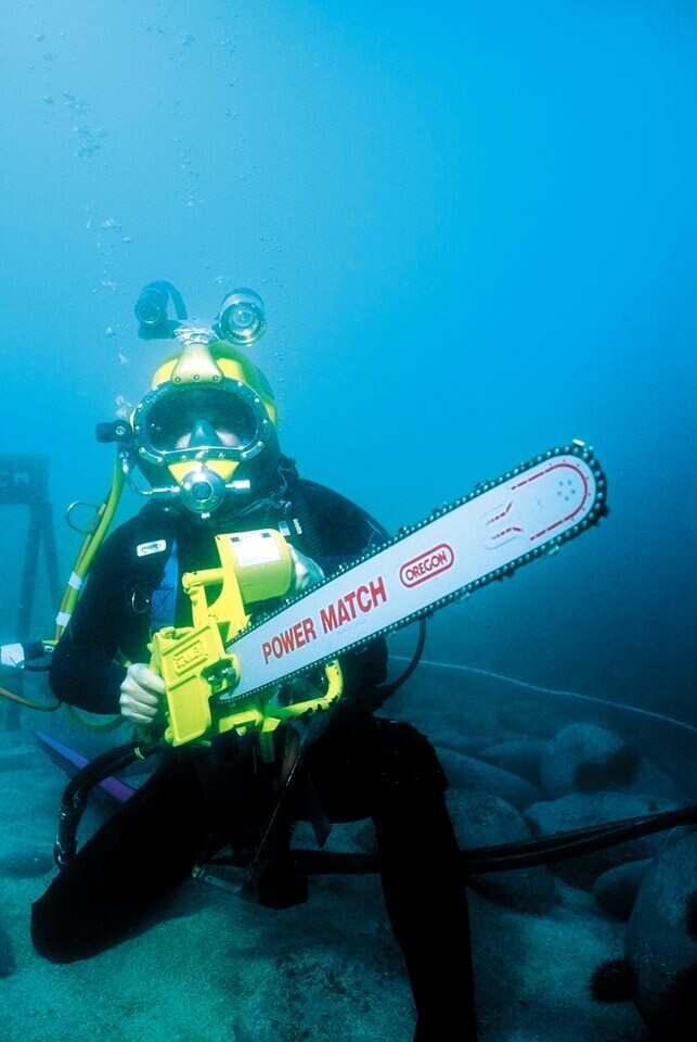 A man underwater in full commercial diving gear, holding a chainsaw.