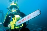 A man underwater in full commercial diving gear, holding a chainsaw.