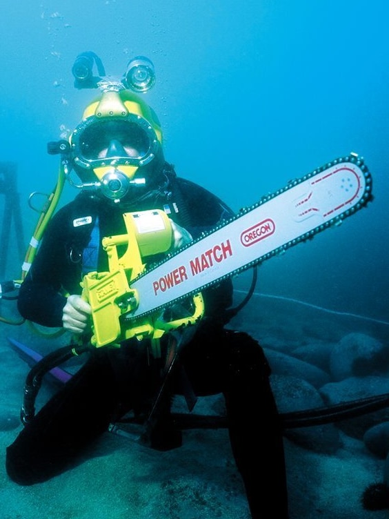 A man underwater in full commercial diving gear, holding a chainsaw.