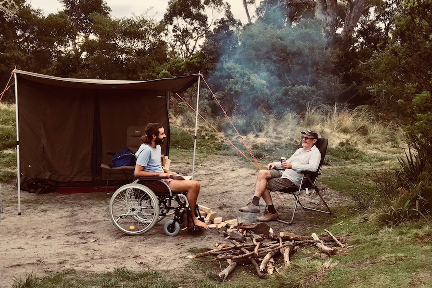 Two men sit at a campsite, one is in a wheelchair the other in a camp chair. They are laughing.
