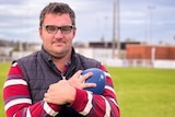 A man wearing glasses standing in a football field holding a football to his chest