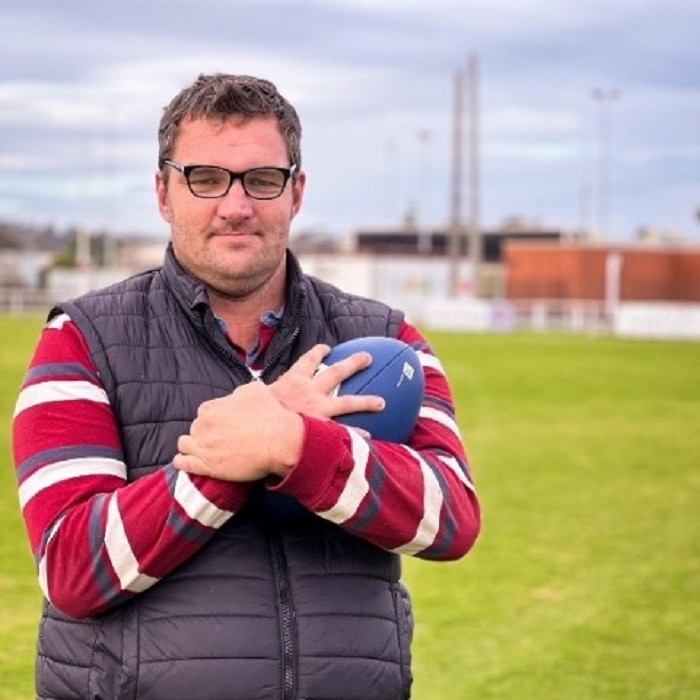 A man wearing glasses standing in a football field holding a football to his chest