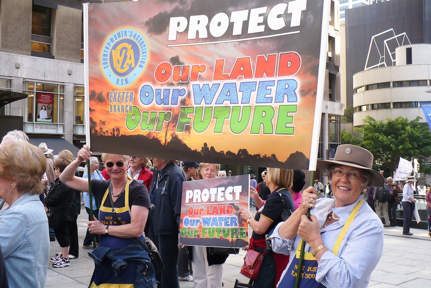 Two ladies wearing CWA aprons stand in a street holding a sign reading 'our land, our water, our future'.