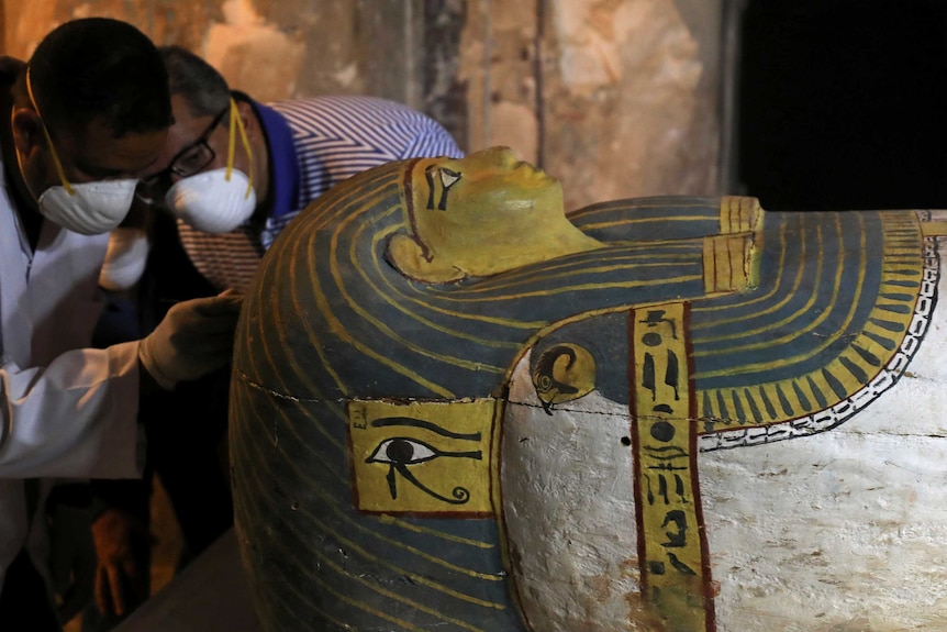 Two men in dust masks examine a large, well-preserved sarcophagus, with bright, clear painting on it