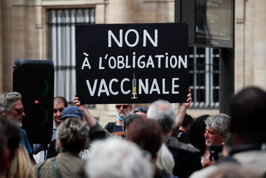 A black and white sign with French words 'Non obligation' is held in tight crowd next to stone-coloured building.