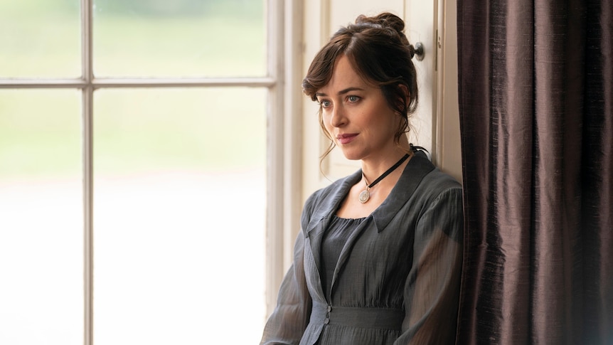 Dakota Johnson sits at a window ledge and looks to the left while wearing a grey dress, with her hair up.