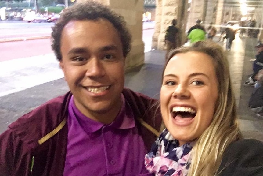 A man wearing a purple shirt and a woman wearing a purple scarf smile while posing in front of a walkway.