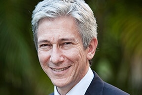 A headshot of a man in a suit smiling. He has grey hair