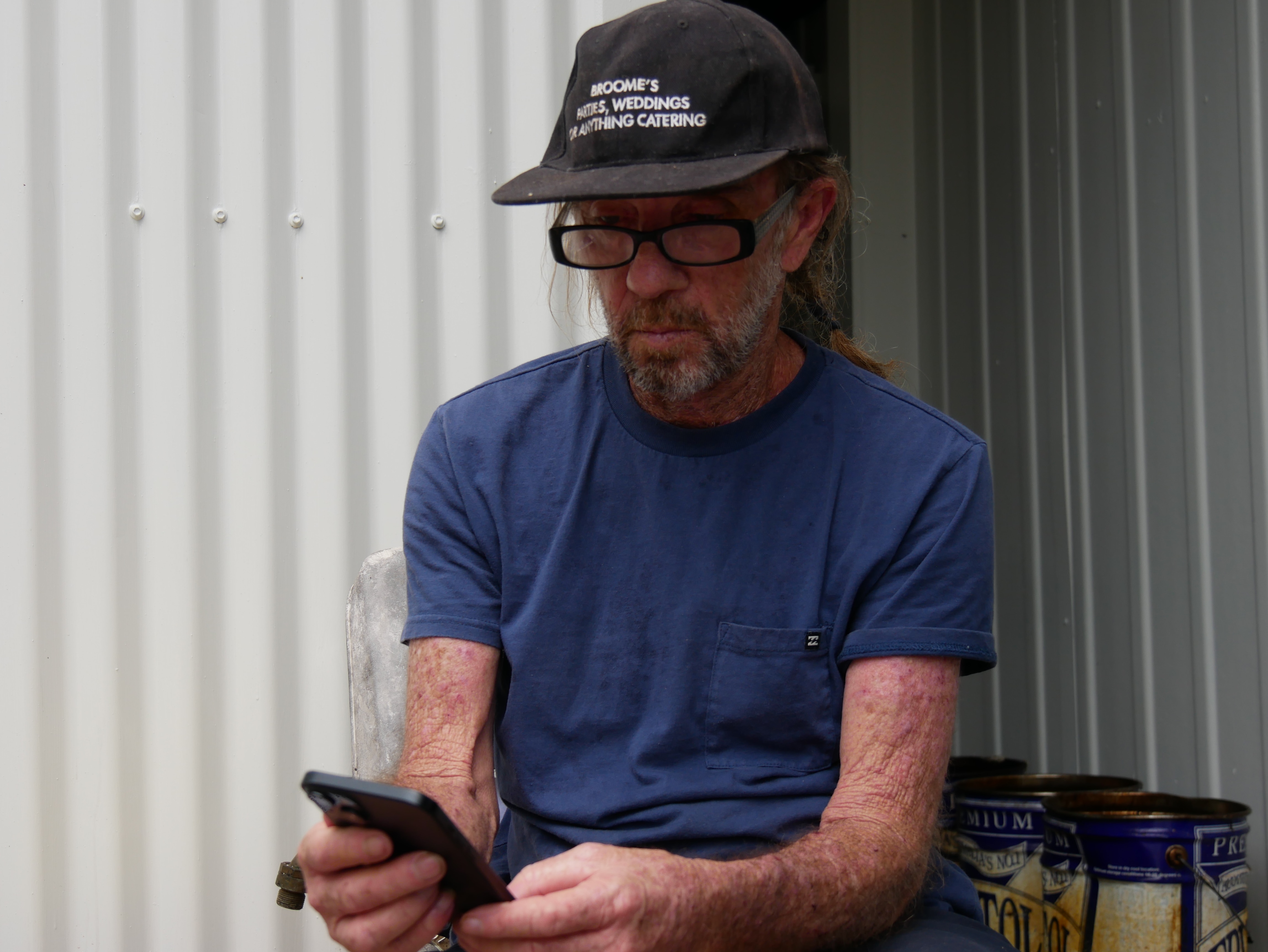 Man with hat, glasses and blue shirt sits while looking down at his phone