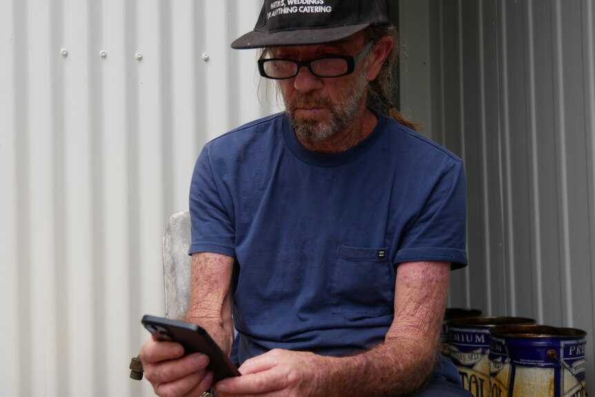Man with hat, glasses and blue shirt sits while looking down at his phone