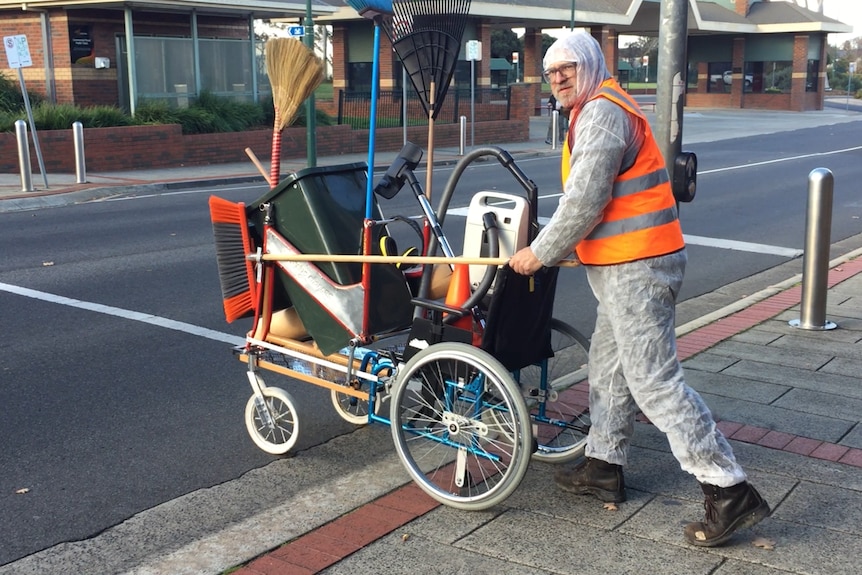 A man in his 50s with glasses and wearing protective clothing pushes a trolley filled with cleaning equipment