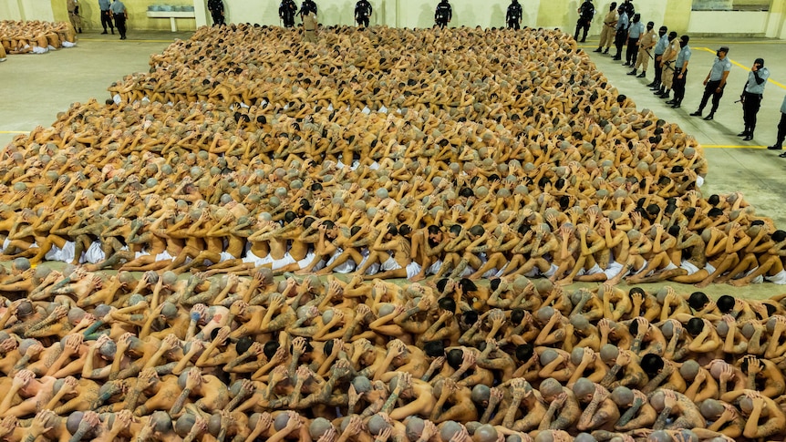 Aerial view of a large group of men sat closely together in rows with their hands on their heads