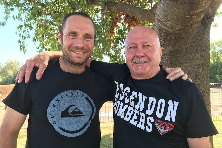 Two smiling men in dark shirts face the camera with their arms around each other.