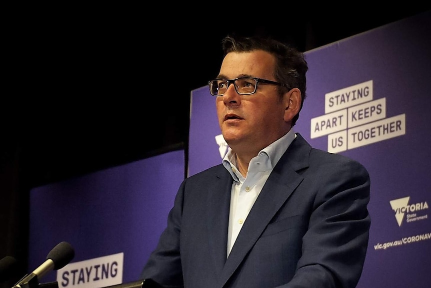 A man in a suit with an open neck collar standing in front of a purple backdrop.