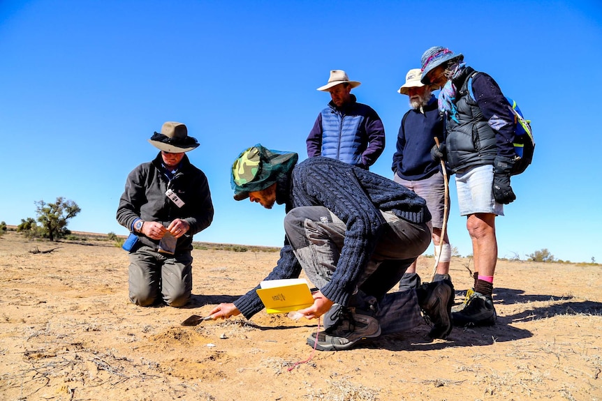 Five people, including two crouching down, survey the desert sand using a trowel.
