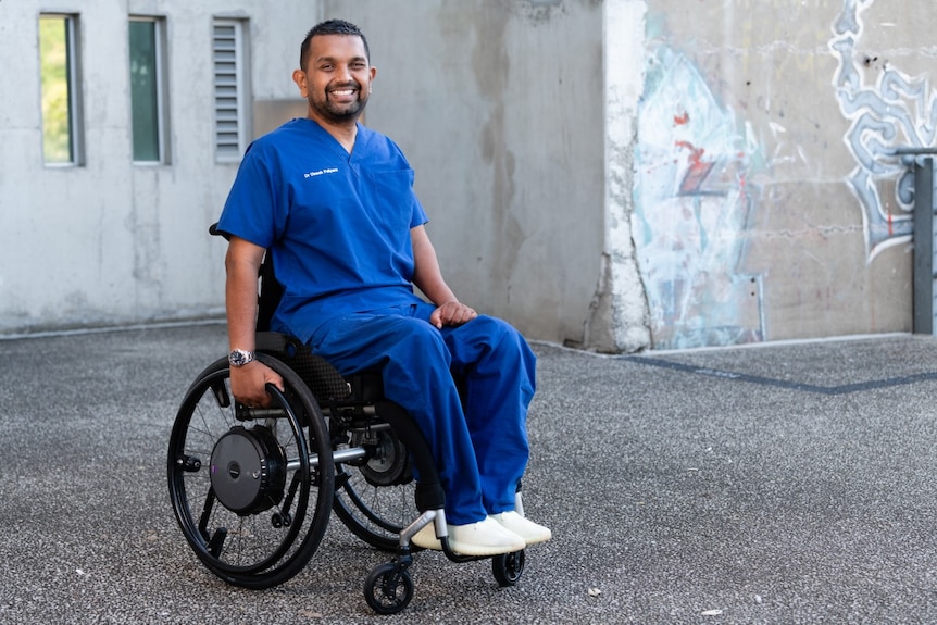 Мъж в сини медицински костюми седи усмихнат в инвалидна количка
