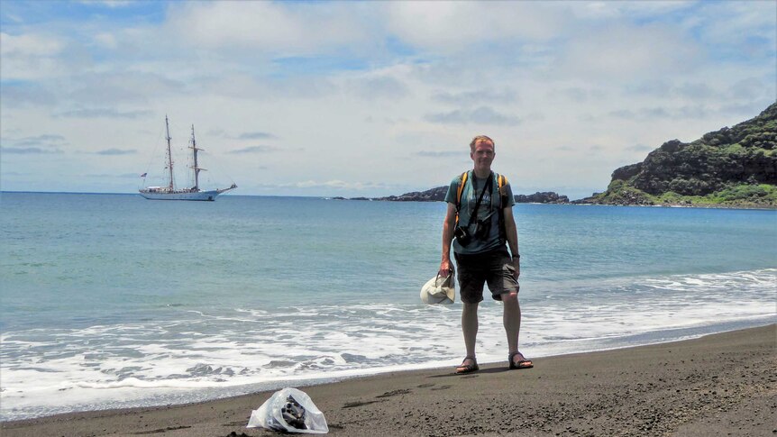 A man in shorts and t-shirt wearing a backpack and camera equipment stands on a black sand beach with a boat in the background
