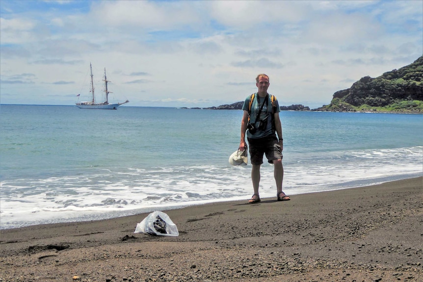 A man in shorts and t-shirt wearing a backpack and camera equipment stands on a black sand beach with a boat in the background