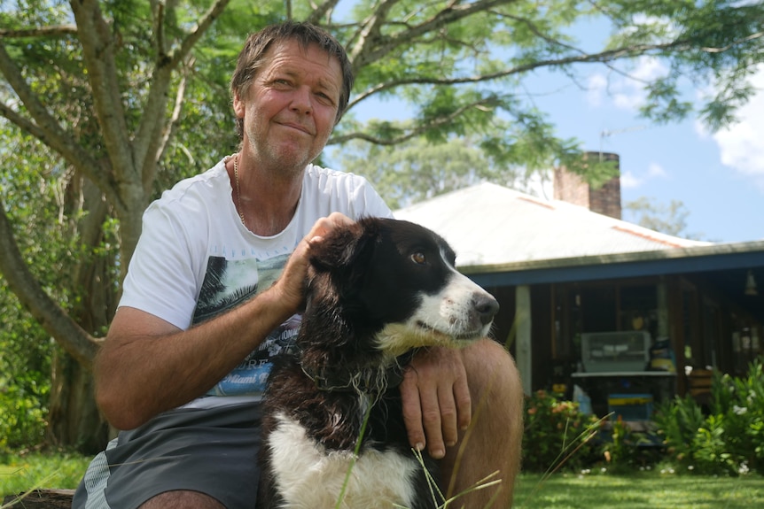 A man in a white tshirt next to a dog with a house in the background.