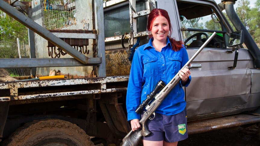 21 year old woman with blue shirt stands in front of a grey utility and holds a grey rifle