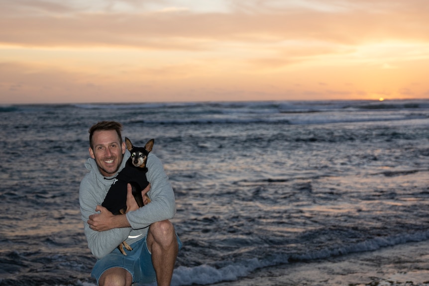 A smiling man holding a small black dog at the beach.