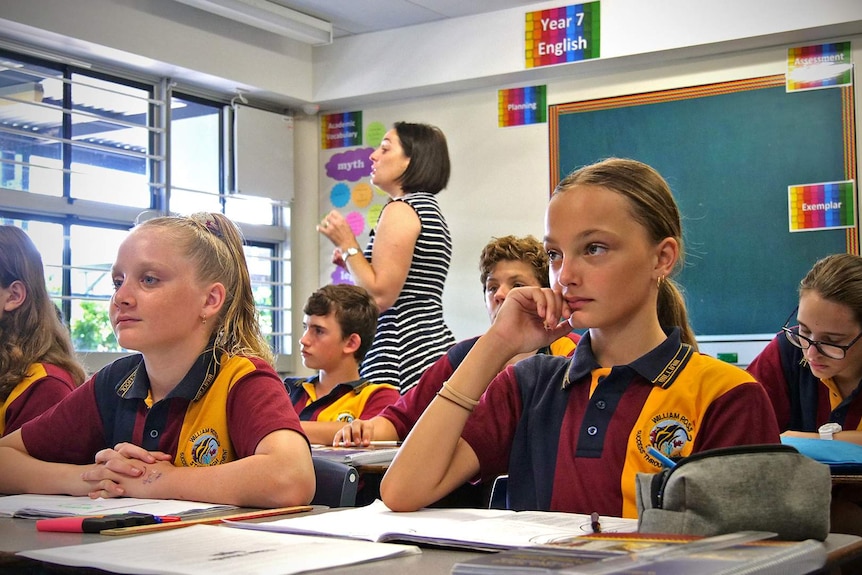 A class of year 7 students sit facing the front while their teacher walks behind them