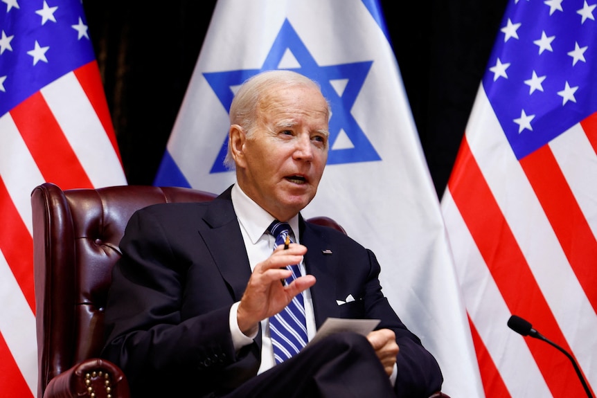 Joe Biden sits in front of Israeli and American flags. He is speaking.