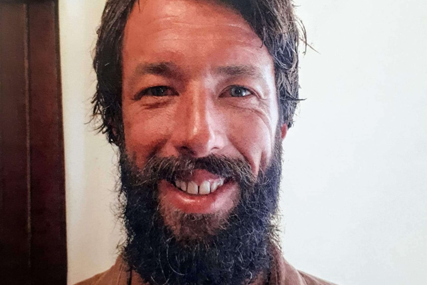 A headshot of a man smiling with a tanned face and long beard.
