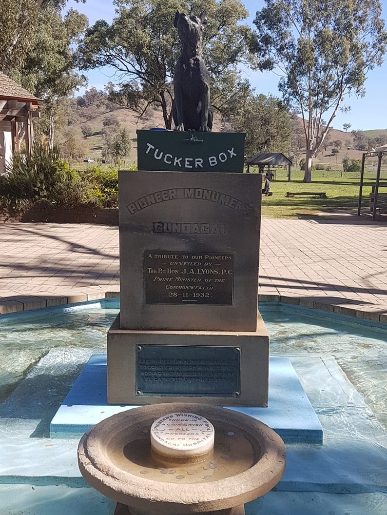 A statue of a dog on a tuckerbox mounted on a plinth in a fountain in a town park.