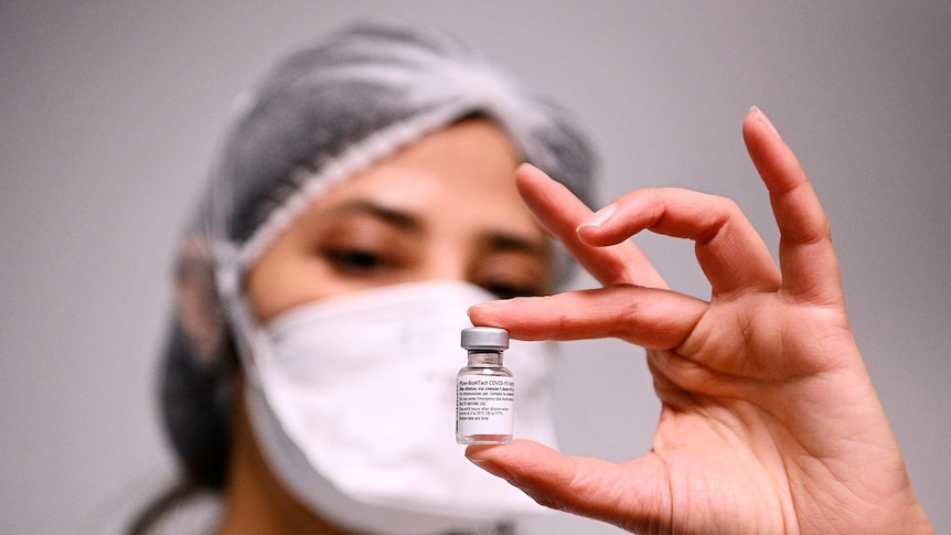 A woman wearing a mask and hair net holds a vial of the COVID-19 vaccine.