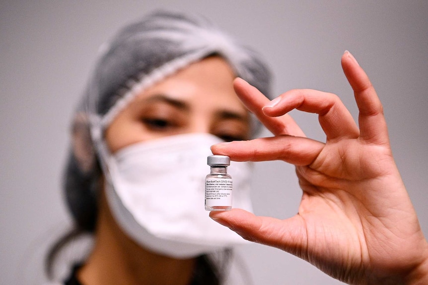 A woman wearing a mask and hair net holds a vial of the COVID-19 vaccine.