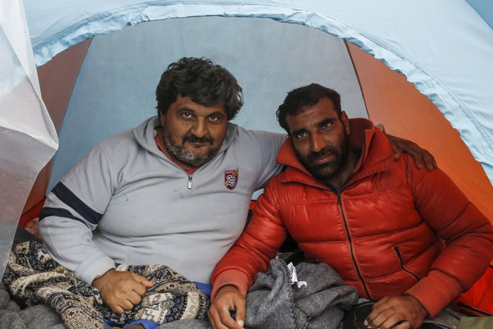 Two men sit inside a tent, one with his arm around the other.