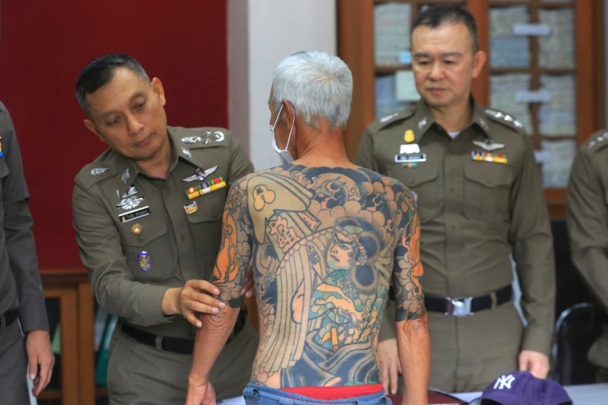 Medium shot of a shirtless man with tattoos being inspected by police.