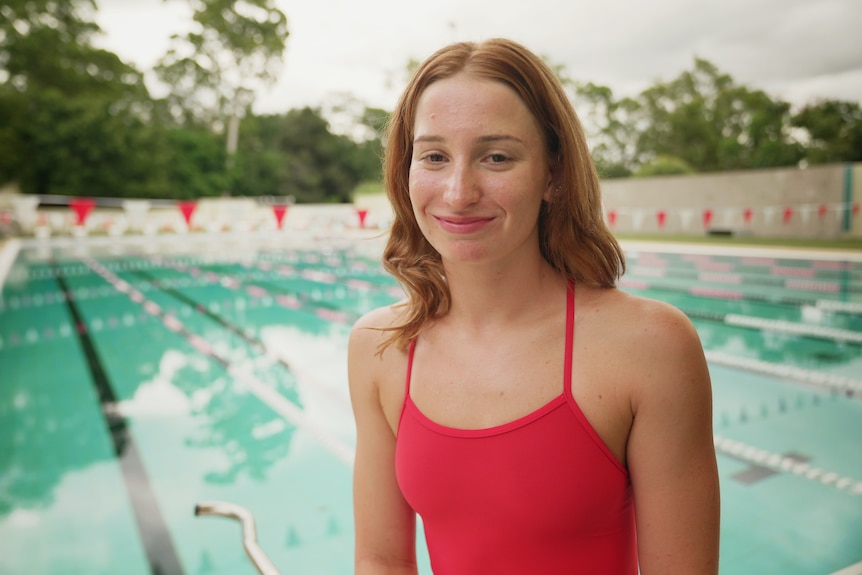 Mollie stands in front of the pool wearing red bathers