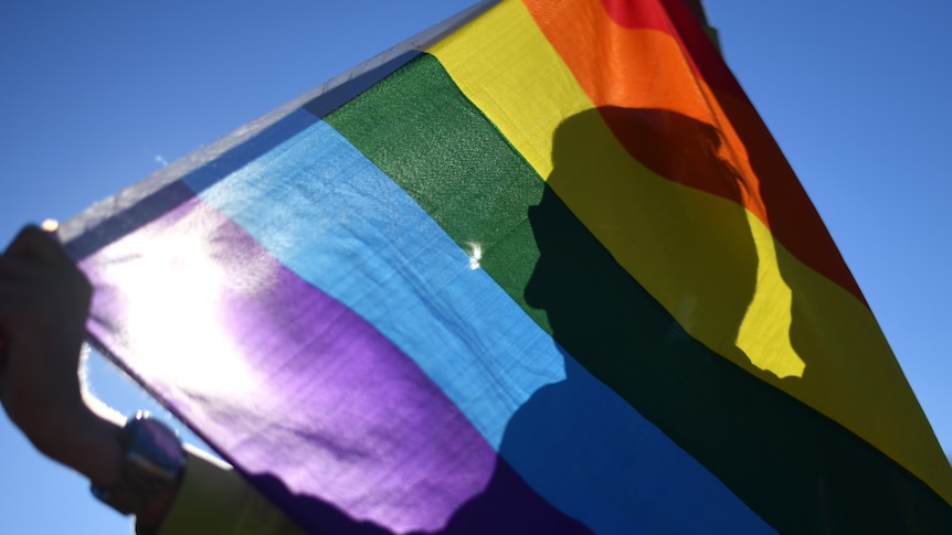 A person in silhouette  holding a rainbow flag on a clear blue sky.