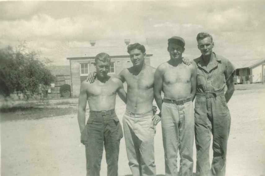 A black and white photo of four young men, three shirtless, with their arms around each other