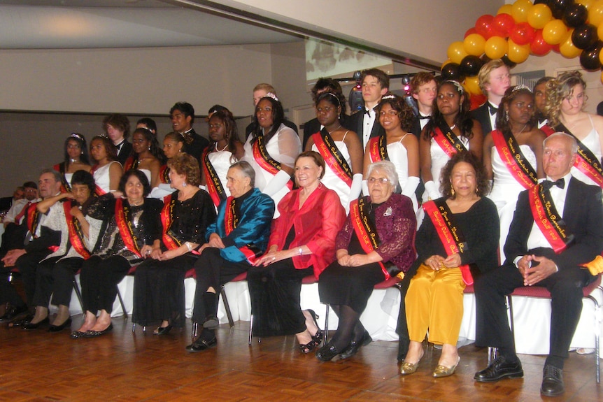 Indigenous women in formal gowns and men in suits stand on tiers behind seated Indigenous elders in a hall.