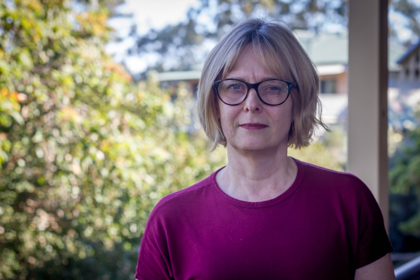 Woman with glasses and purple sweater stands on a porch.