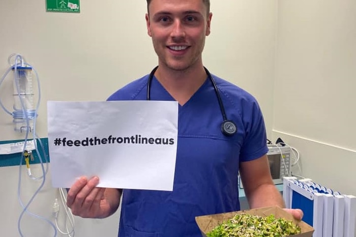 A smiling man with a stethoscope holds a bowl of food and a sheet of paper printed with hashtag feed the frontline aus
