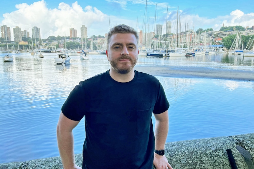 A man in a black t-shirt is pictured in front of boats moored on the water.