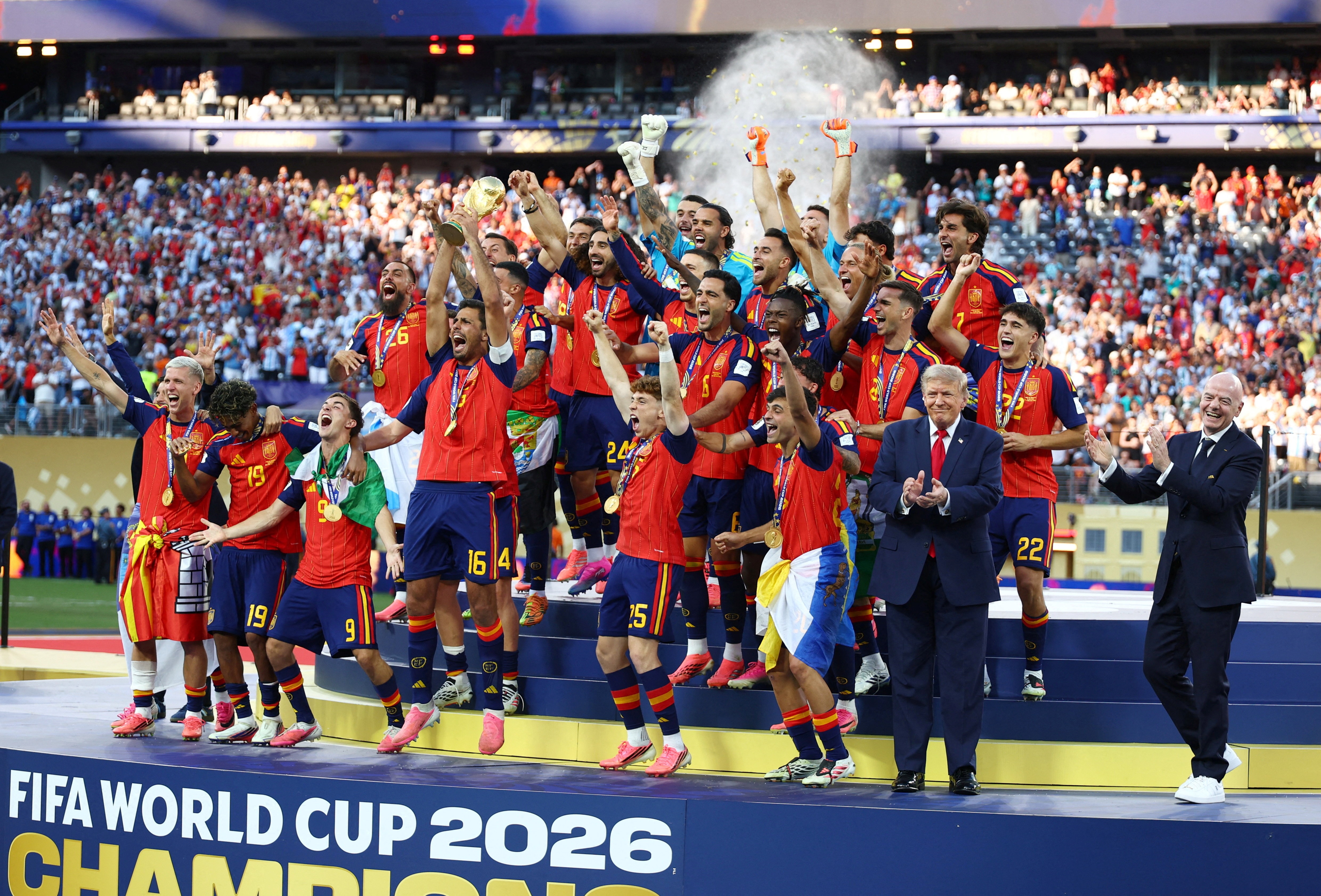Men in suits applaud while footballers celebrate on a podium in a packed stadium.