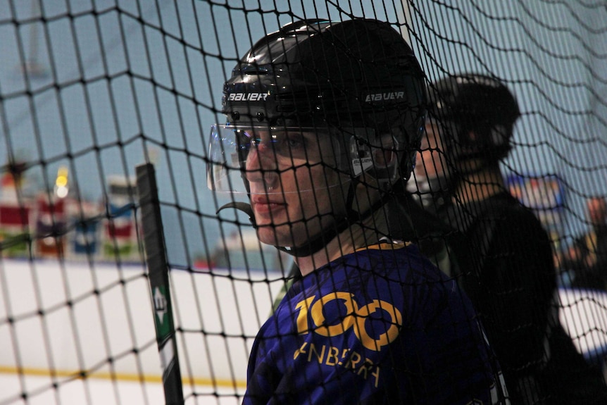 A man stands at the side of the ice skating rink watching his team mates.