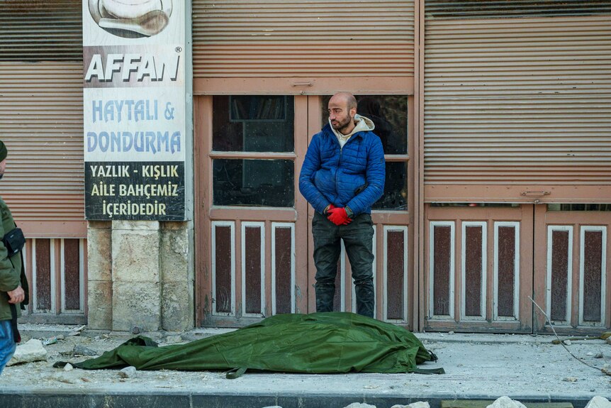 A man stands next to a green tarp covering up a body 