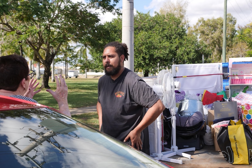 A man stands in front of a trailer full of bags and furniture and talks to a woman in front of her car