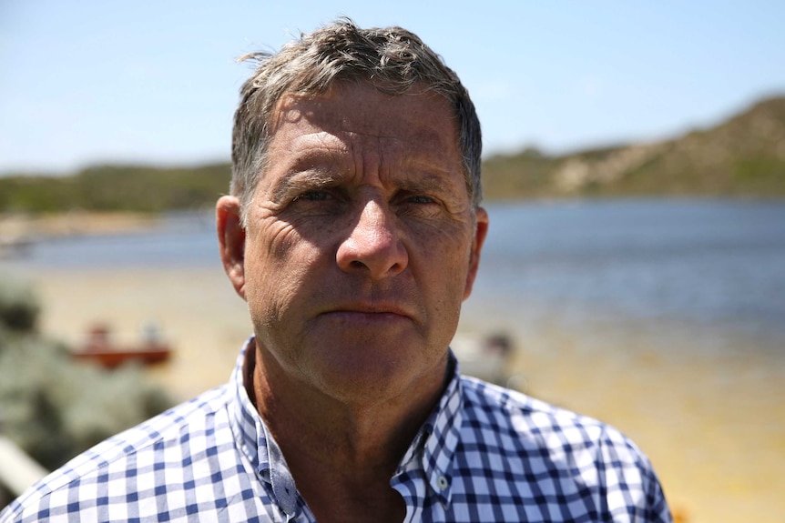 A headshot of a man in a blue and white checked shirt in front of a river.