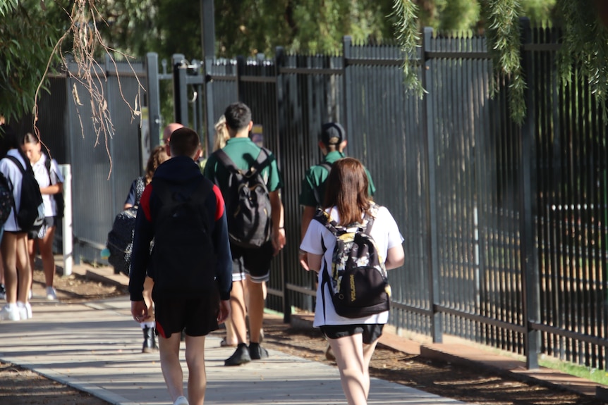 Teenagers in white and black school uniforms walk along a black fence.