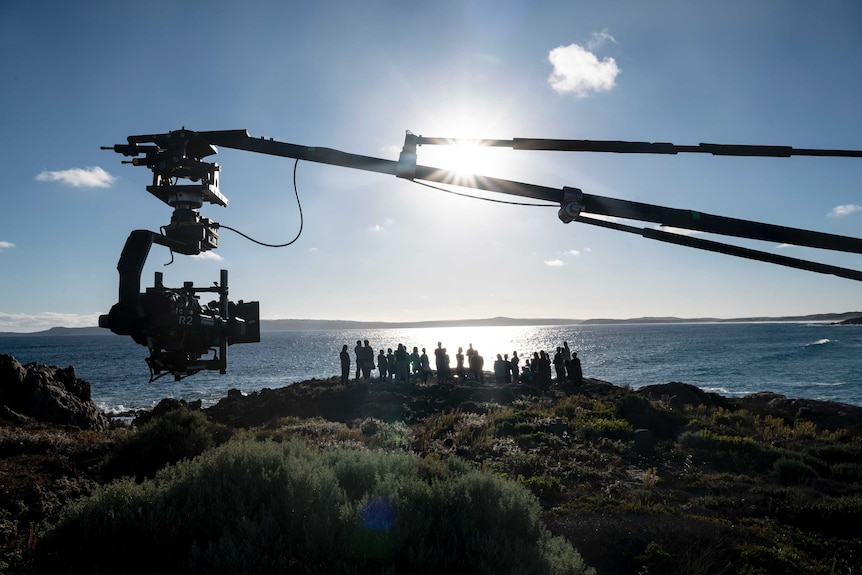 A group stands on a coastal setting on a sunny day. Blue sky, looking into the sun. Camera boom is visible.