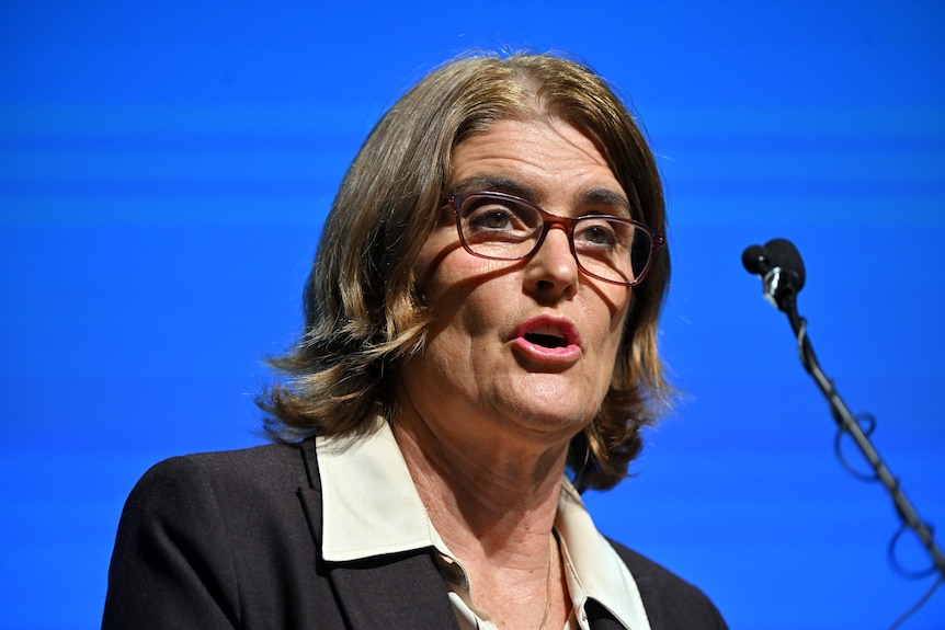 An older woman with a dark grey bob and glasses speaks into a small microphone in front of a blue screen.