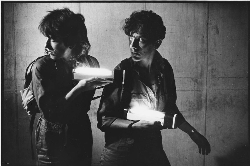 Black and white: two women peer behind them while holding glowing boxes.