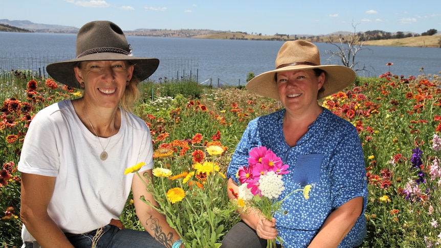 Two women kneel in a field of colourful flowers with a lake behind them 